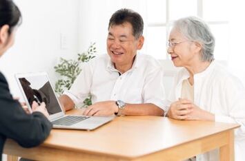 A real estate agent showing a laptop to a senior couple at a dining table. シニア夫婦にラップトップで説明する不動産エージェント。