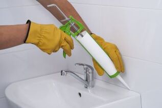 Yellow gloved hands holding a caulking gun caulking around the sink in the bathroom.