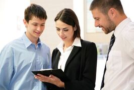 2 men are looking at the clip board the woman in the middle is holding in the business setting.