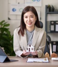 Young Asian woman in khaki blazer holding a house in her hands at office desk.
