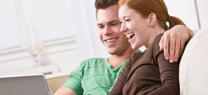 A young couple smiling looking at a laptop. Man wearing a green shirt and his arm is placed around her shoulder. Woman with pony tail wearing a brown shirt.