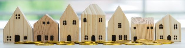 7 different wooden homes lined up on a table with gold coins scattered in front.